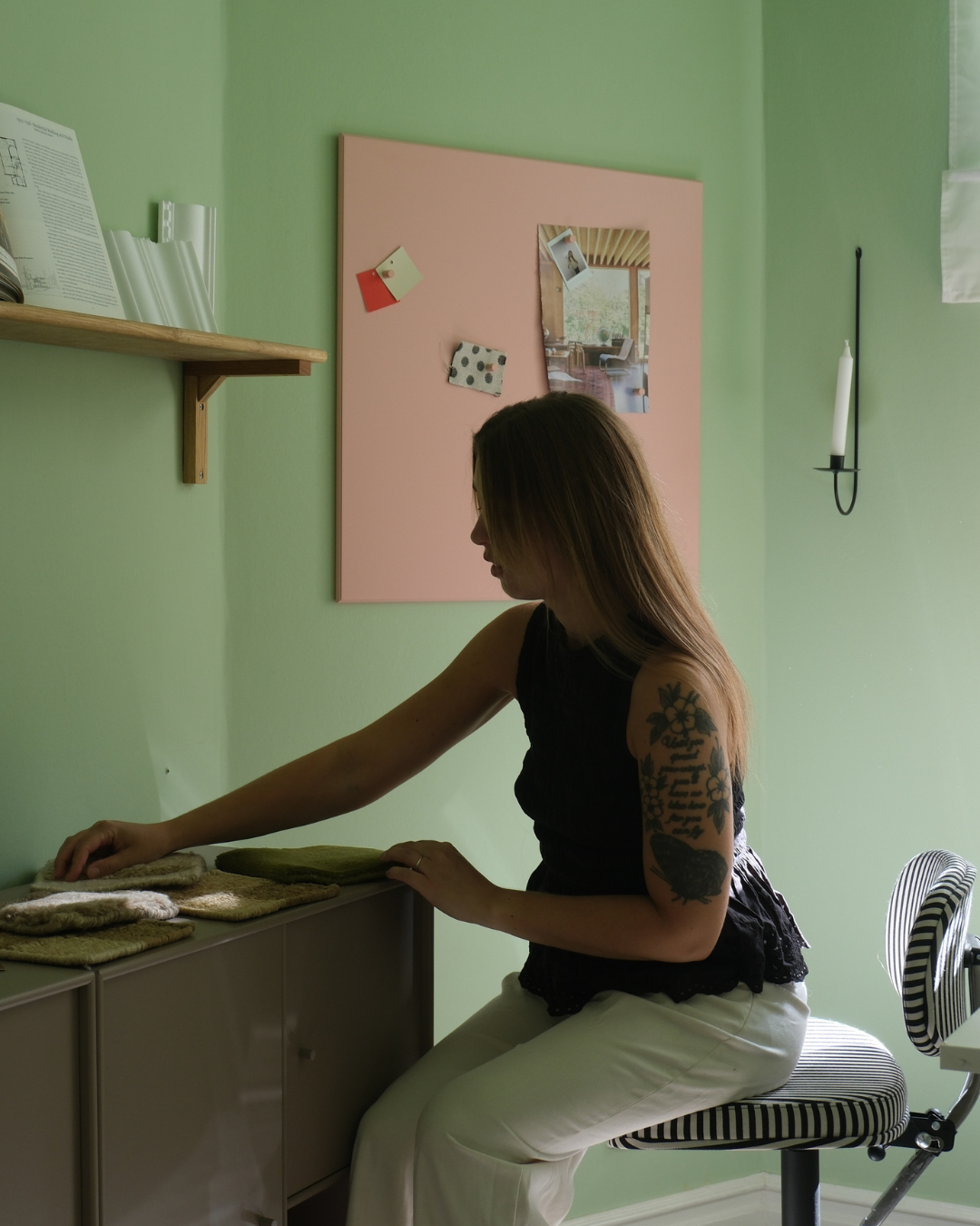A woman with a tattoo on her left arm sitting on a striped office chair, reaching towards a collection of fabric swatches on a gray cabinet in a room with green walls.