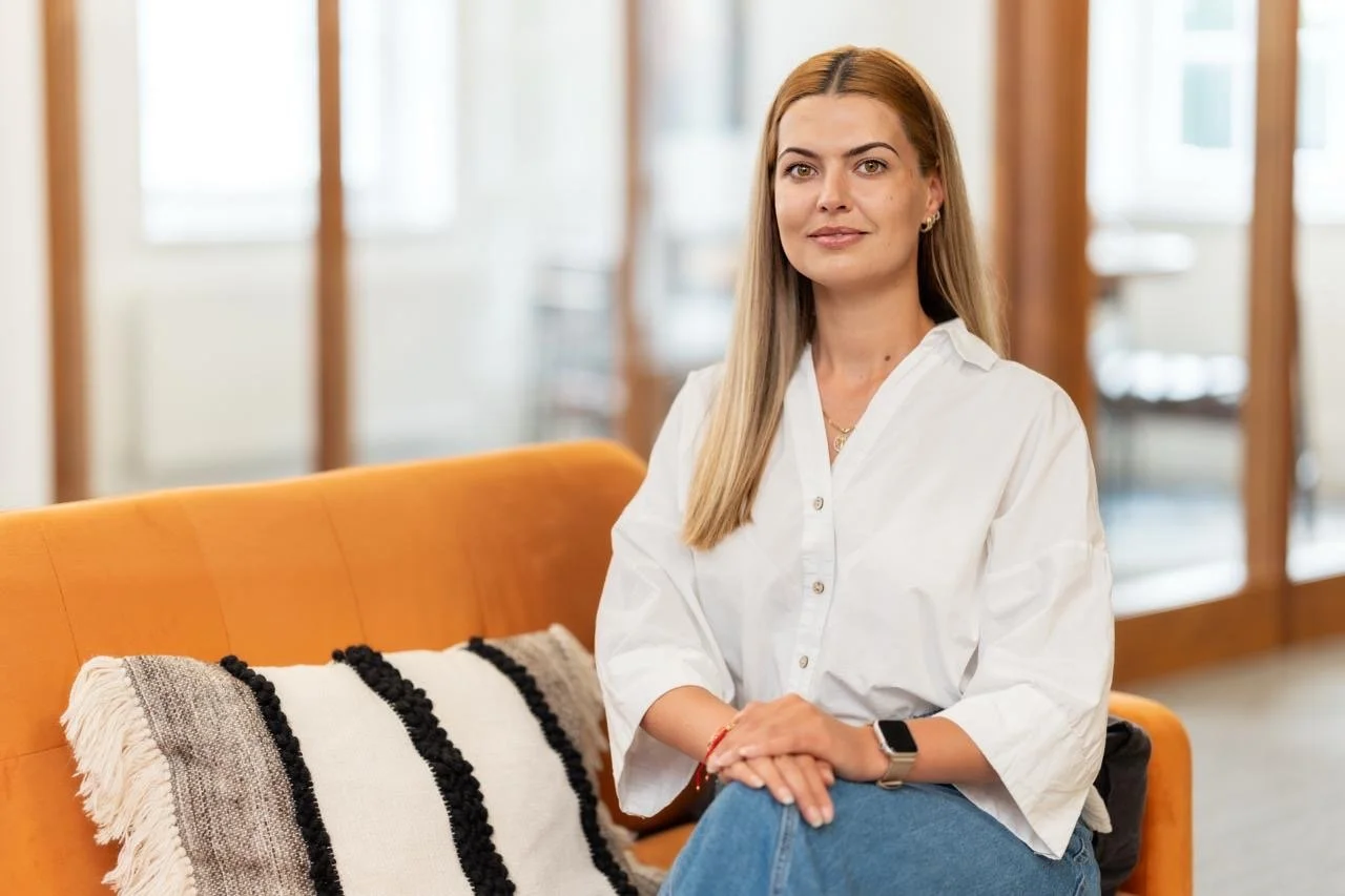 Vladislava Yankova, Founder and Director of Ledger Leaf Ltd. The image shows a woman with long hair wearing a white button-up shirt and jeans, sitting on an orange sofa with a black and white pillow, in a bright room with large windows.