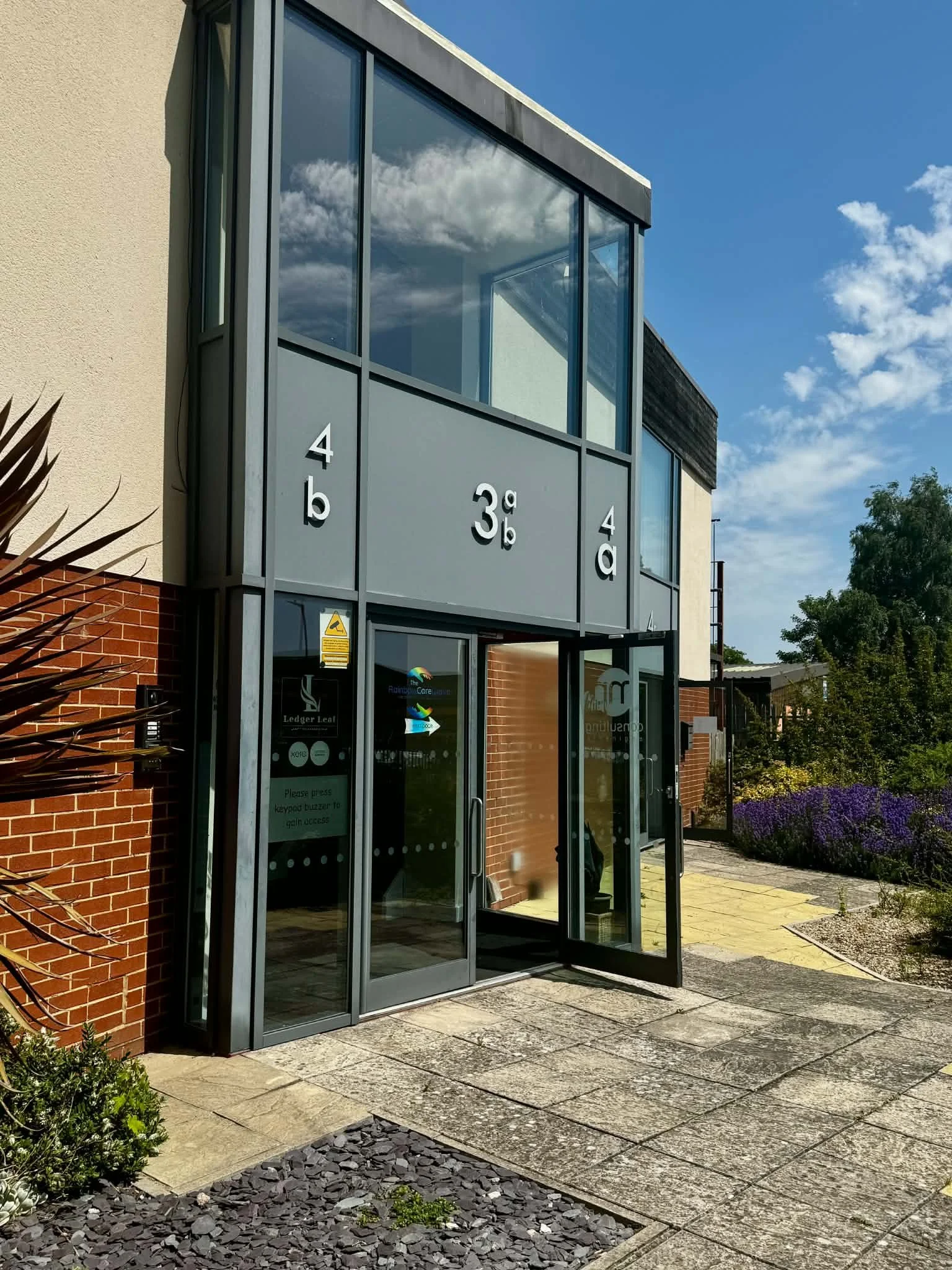 Exterior view of a modern building with glass windows and brick wall, featuring numbered signs on the glass entrance, clear sky, and landscaped walkway with plants and gravel.