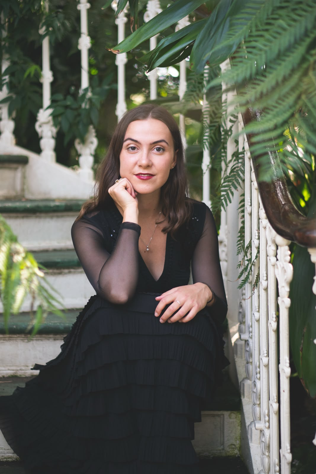 A woman sitting on outdoor stairs surrounded by lush greenery and white ornate railing, wearing a black dress with sheer long sleeves, looking at the camera with a slight smile.
