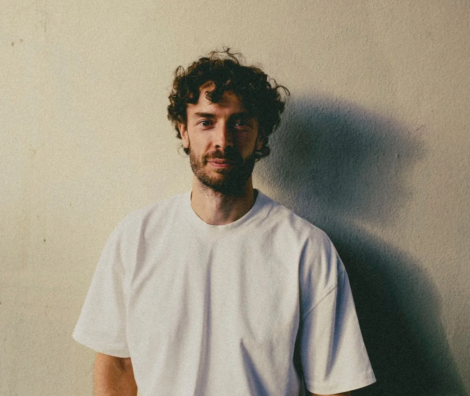 A young man with curly dark hair and a beard, wearing a white t-shirt, standing against a plain wall with a shadow cast on the right side.