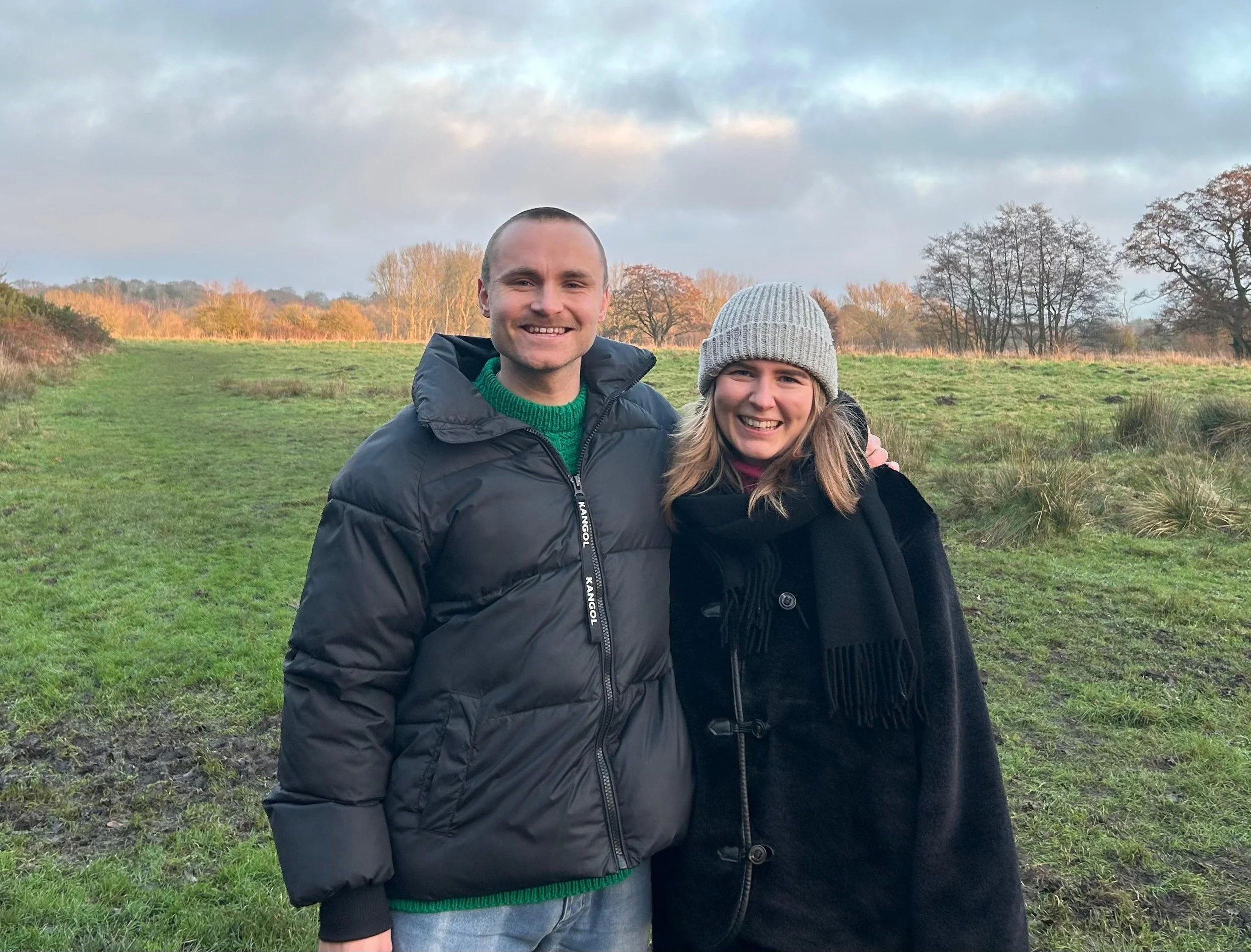 A man and a woman smiling outdoors in a grassy field with trees and a cloudy sky in the background, wearing warm clothing.