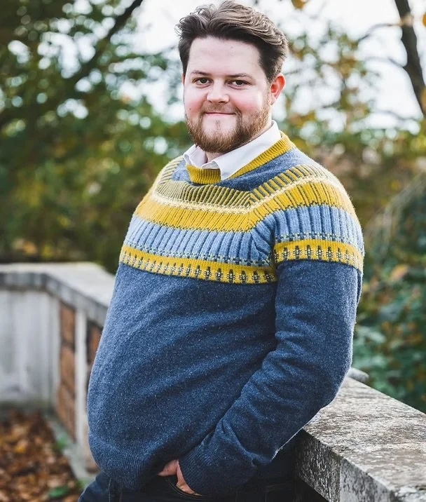A young man with dark hair and a beard, wearing a colorful striped sweater, stands outdoors in front of a wooden railing and trees with green leaves.