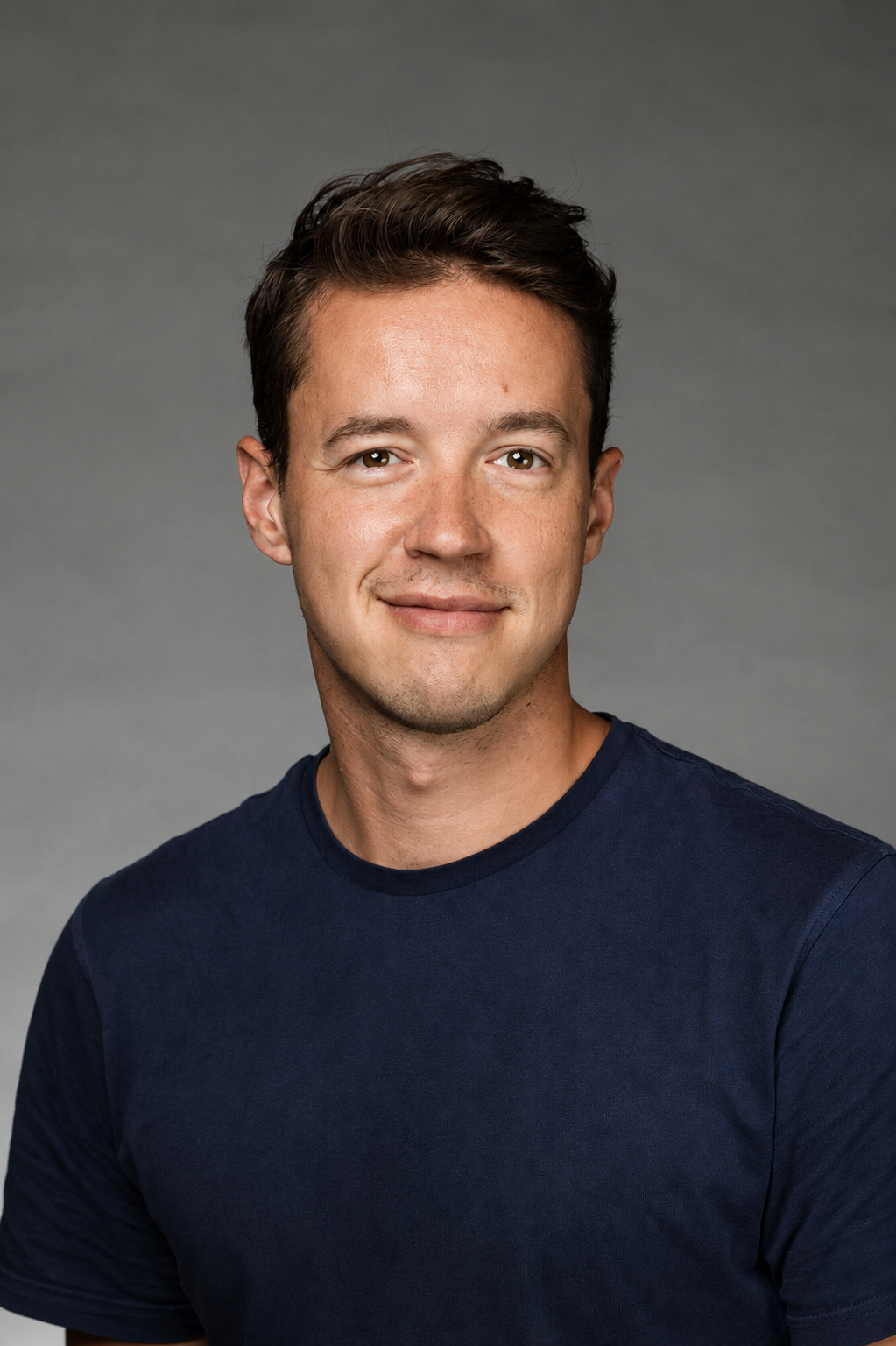 Headshot of a young man with short brown hair, wearing a navy blue t-shirt, smiling softly against a neutral gray background.
