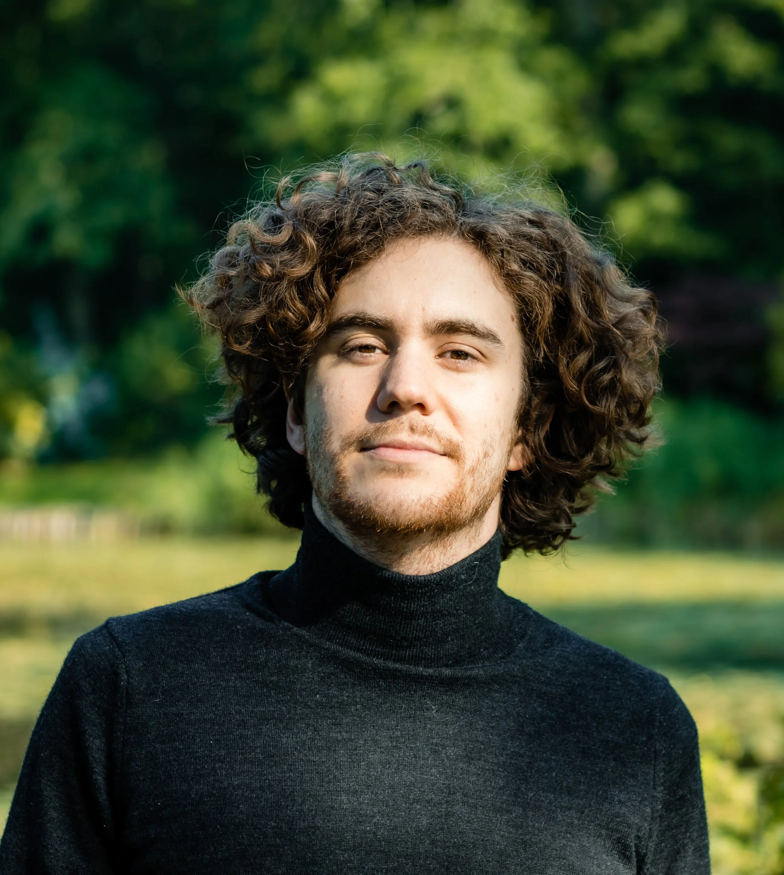 Close-up of a young man with curly hair and a beard, wearing a black turtleneck sweater, outdoors with green trees and grass in the background.