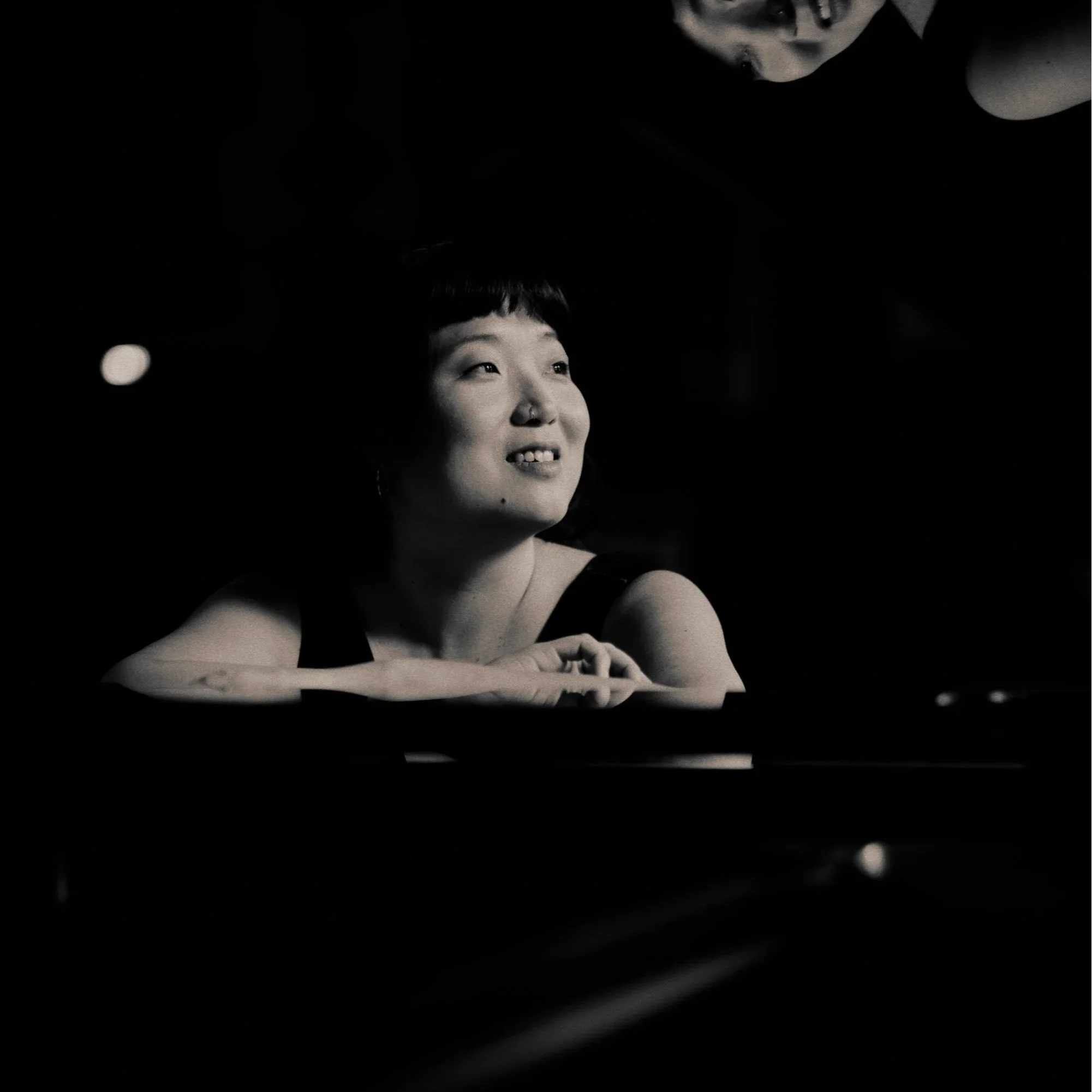A black-and-white photo of two women in a close, intimate pose, one gazing upward with a slight smile and the other looking down, with a dark background and soft lighting highlighting their faces and arms.