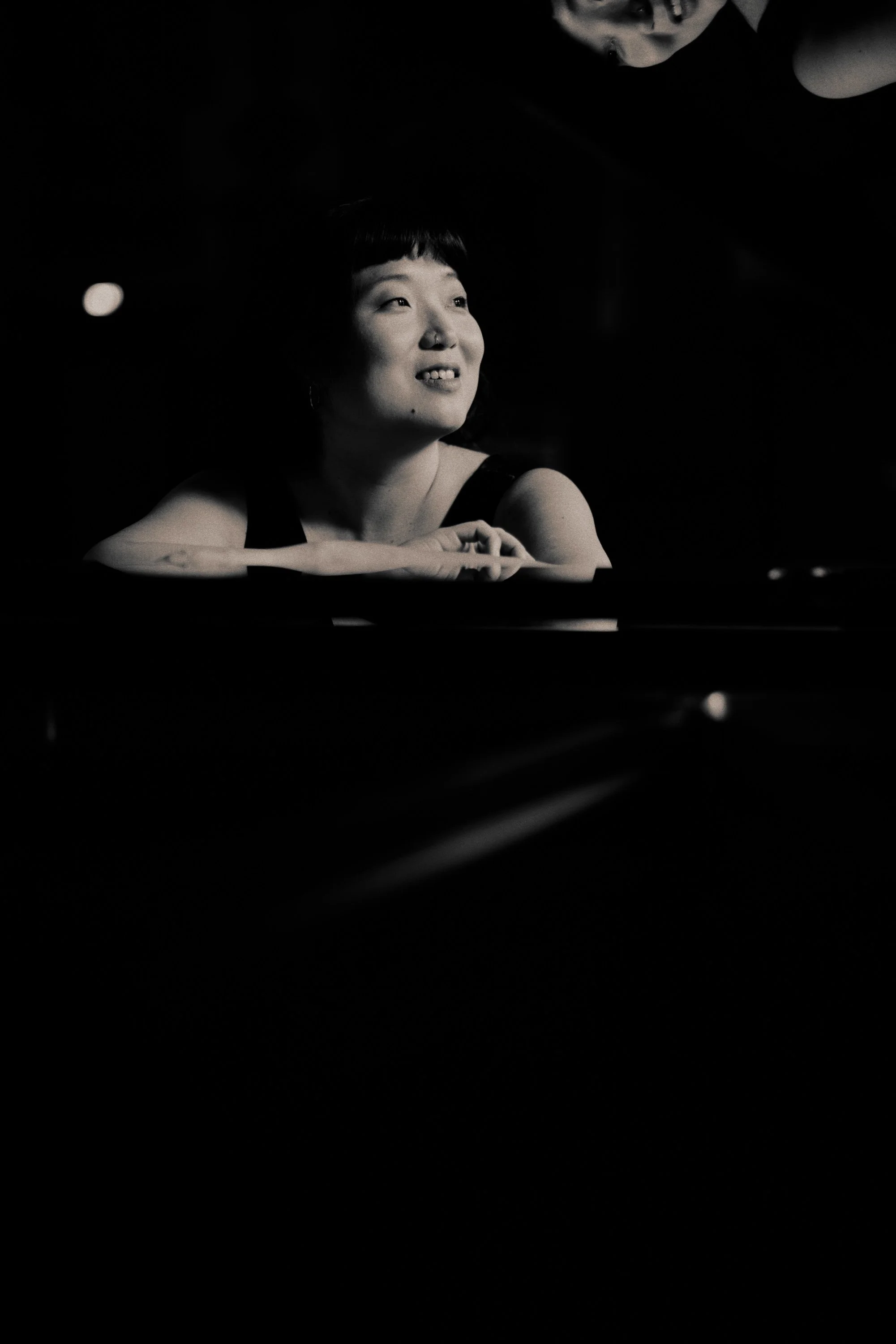 A black and white photo of a woman with short hair, looking slightly upward and smiling, with a person's face partially visible above and to the right.