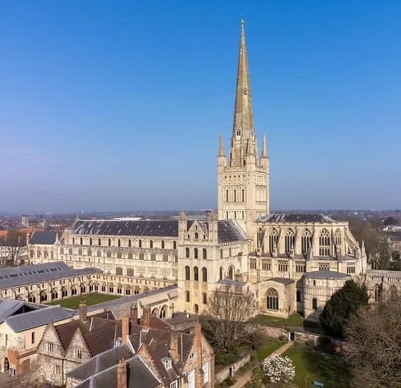 A large historic church with a tall spire and Gothic architecture, surrounded by smaller buildings and a garden under a clear blue sky.