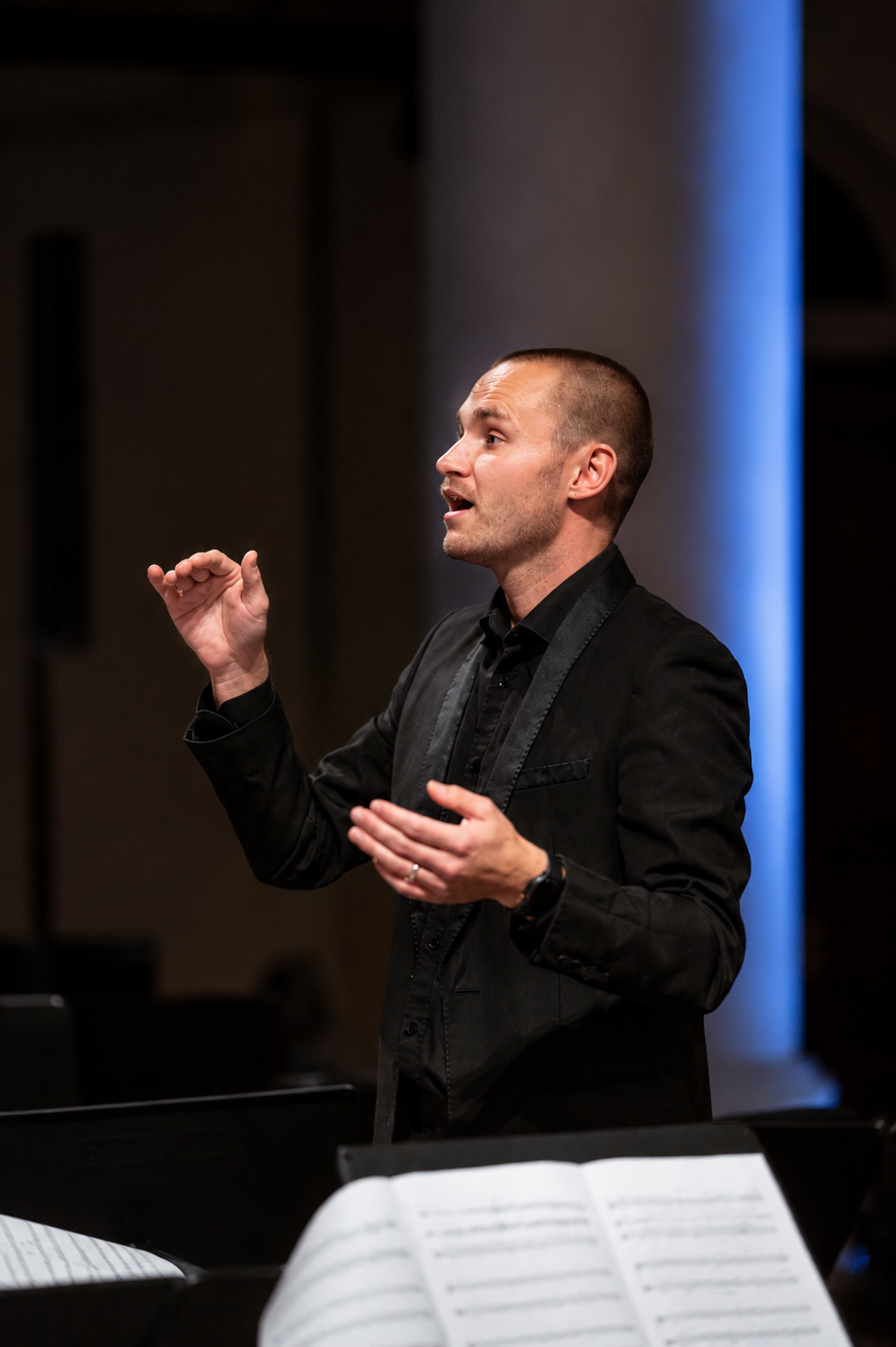 A man with short hair, wearing a black suit and shirt, conducts an orchestra in a dimly lit concert hall, with sheet music visible in the foreground.