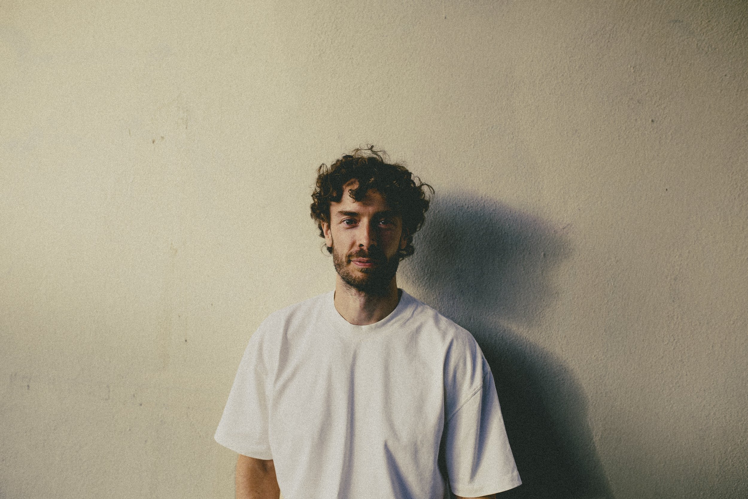 A young man with curly hair and a beard wearing a white t-shirt, standing against a plain beige wall.