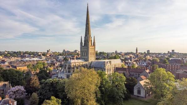 A large Gothic-style church with a tall steeple in a cityscape, surrounded by trees and smaller buildings, under a partly cloudy sky.
