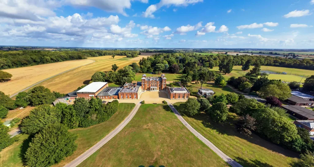 A large historic mansion with red brick walls and white accents, surrounded by trees and open fields under a partly cloudy sky.