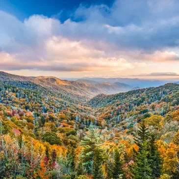 A scenic view of a colorful autumn landscape with rolling hills covered in trees with fall foliage under a partly cloudy sky.
