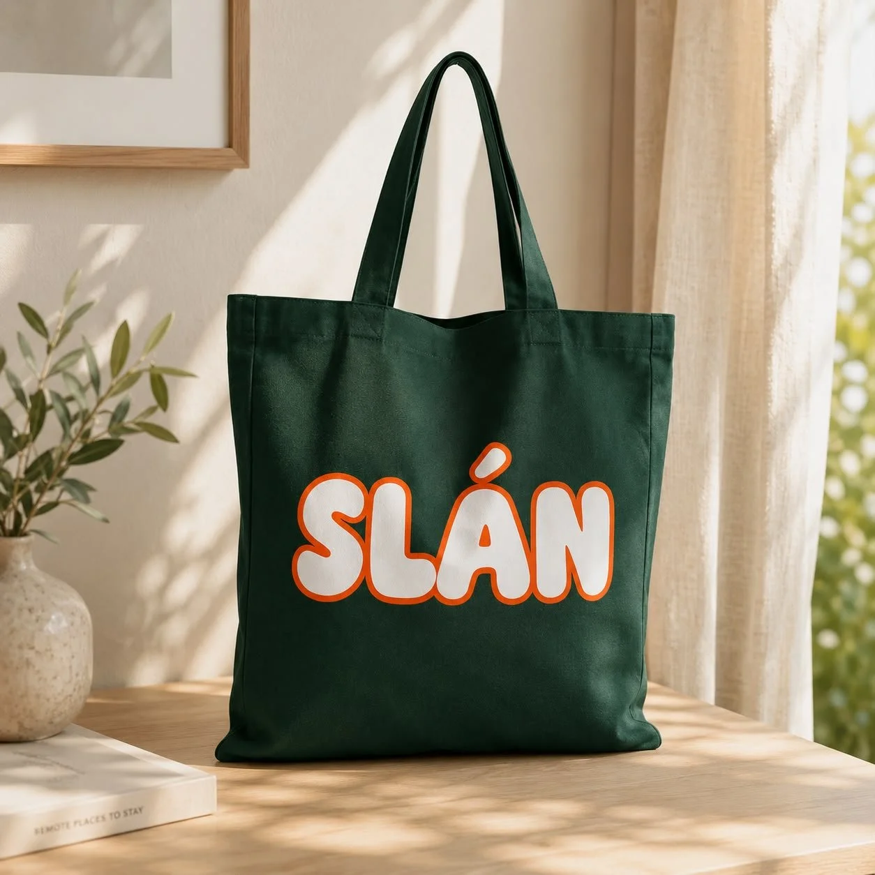 A green tote bag with the word "SLÁN" in large white and orange letters, hanging inside a room near beige curtains and a potted plant on a wooden table.