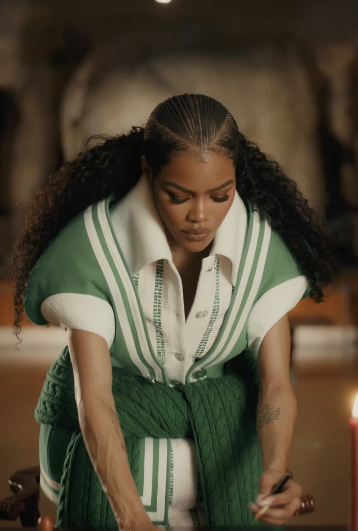 A woman with long curly hair, wearing a green and white striped shirt and a green knit sweater tied around her waist, is looking down with a thoughtful expression in a warmly lit room.
