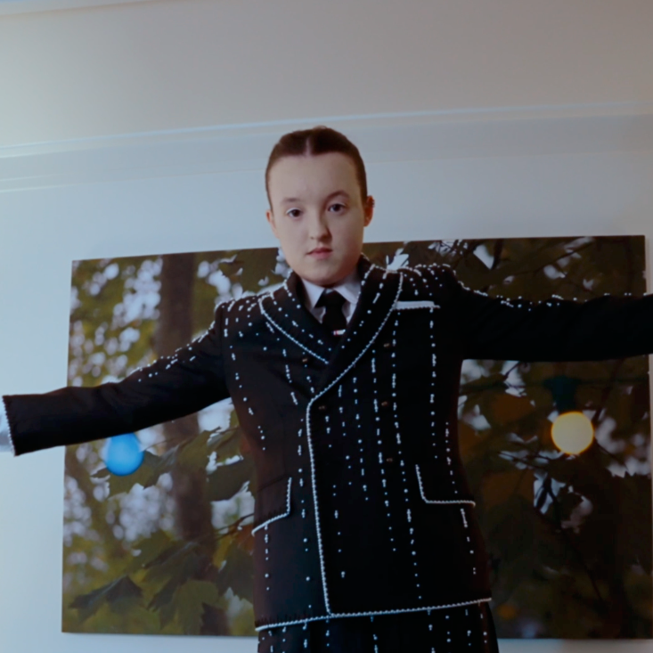Girl in a black blazer with white stitching, standing with arms outstretched in front of a nature background.