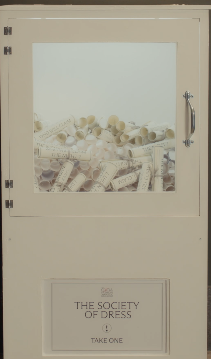 A white donation box with a glass window filled with rolled paper tickets labeled "Who Will Claim the Night?" and a sign below that reads "The Society of Dress - Take One."
