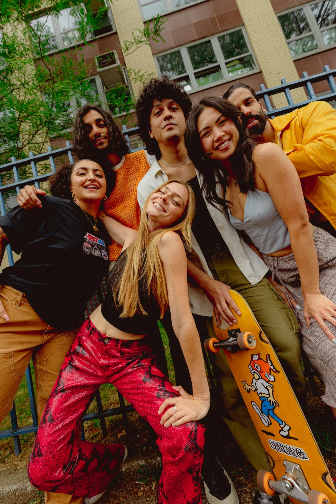 Group of six diverse young people outdoors behind a metal fence, smiling and posing with one person holding a skateboard featuring cartoon animals, a building with multiple windows and trees in the background.
