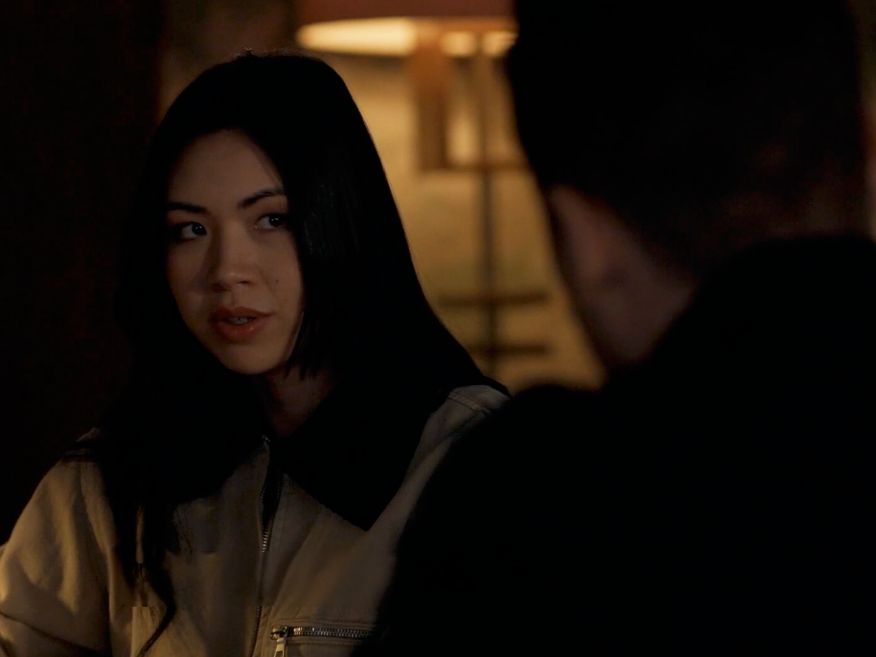 A young woman with long black hair and a light-colored jacket looking at a person with short dark hair in a dark room with warm lighting.
