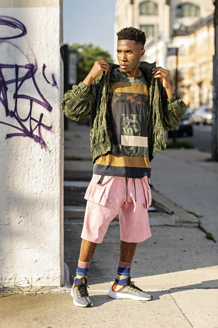 Young man standing on a city sidewalk near a graffiti-covered wall, adjusting his green bomber jacket while wearing a graphic t-shirt, pink shorts, colorful socks, and running shoes.