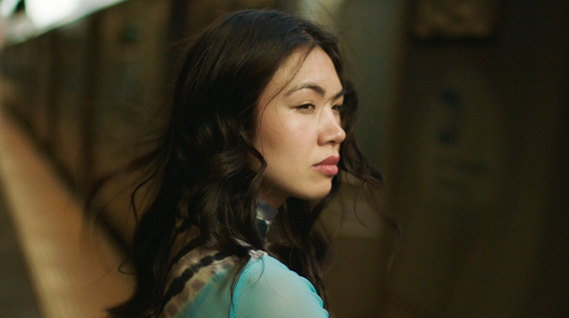 A young woman with dark, wavy hair looking to the side, wearing a light blue top, with a neutral expression, in an indoor setting.