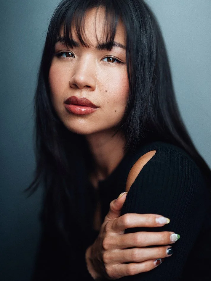 A woman with shoulder-length black hair and bangs, wearing a black top with a shoulder cutout, posing against a neutral background.