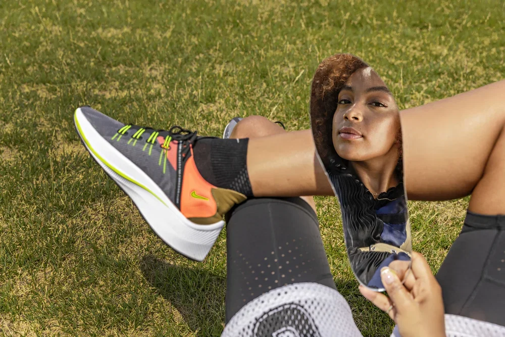 A woman with curly hair stretching outdoors on the grass, wearing athletic clothing, near a black foam roller and a running shoe.