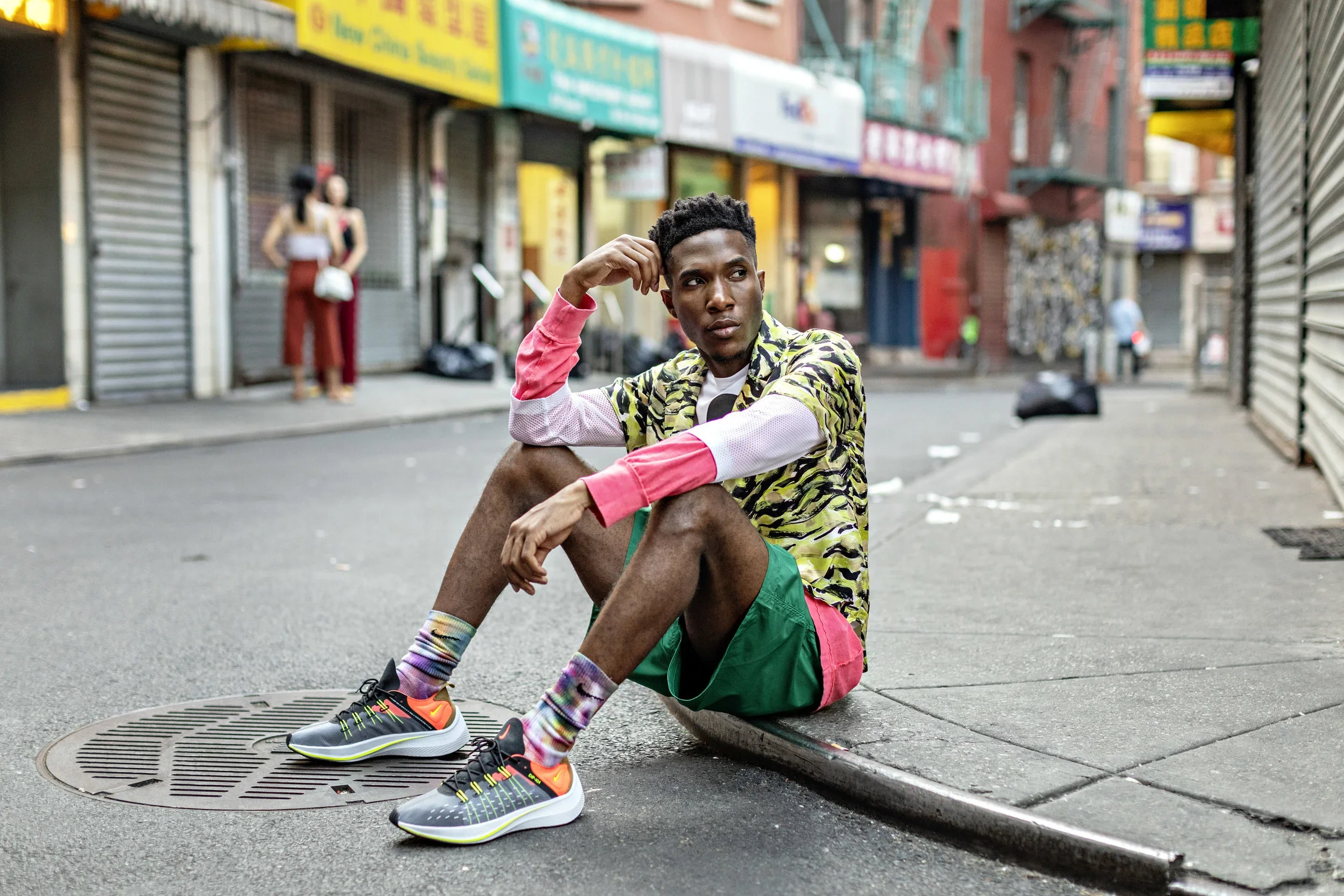 A young man wearing colorful sneakers, tie-dye socks, and bright clothing sits on a city street curb near a manhole cover, with two women in the background and storefronts across the street.