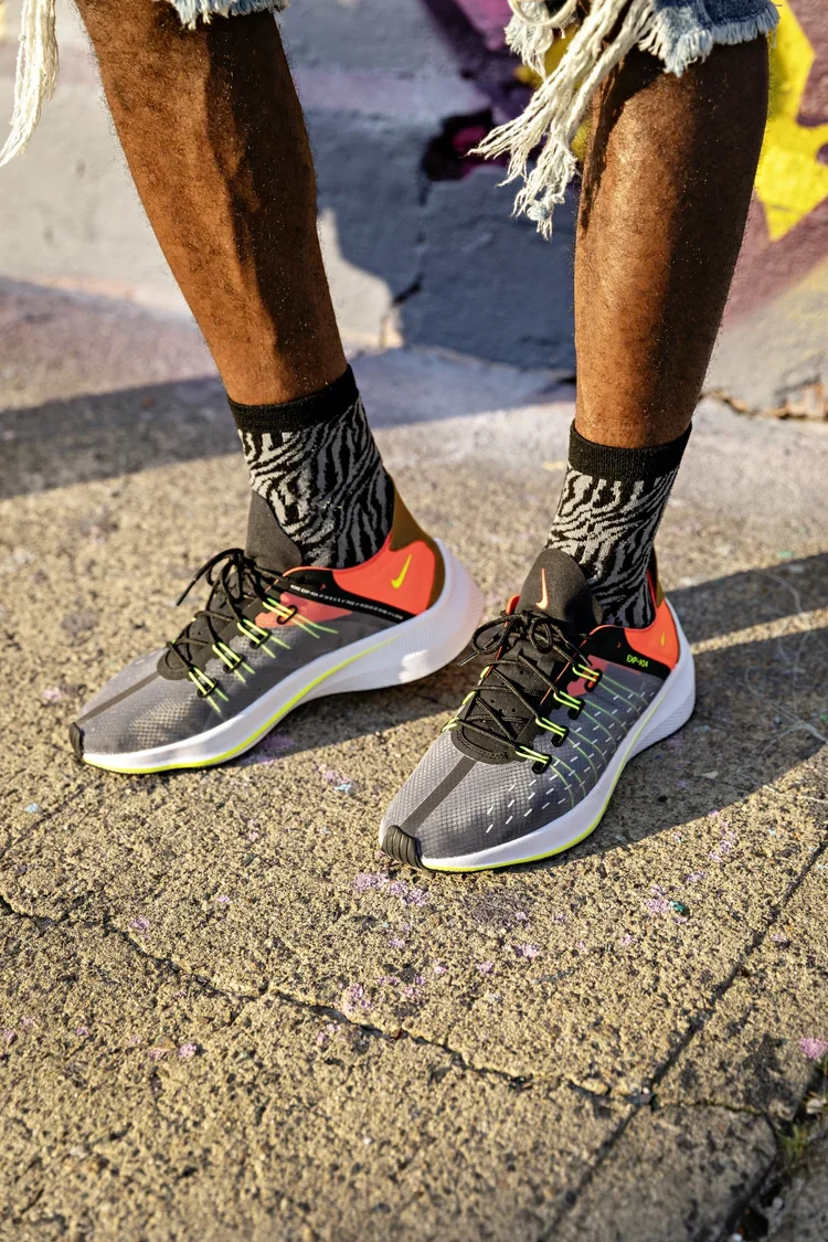 Close-up of person's feet wearing black and gray Nike running shoes, zebra-print socks, and torn denim shorts, standing on a sidewalk with sunlight casting shadows.
