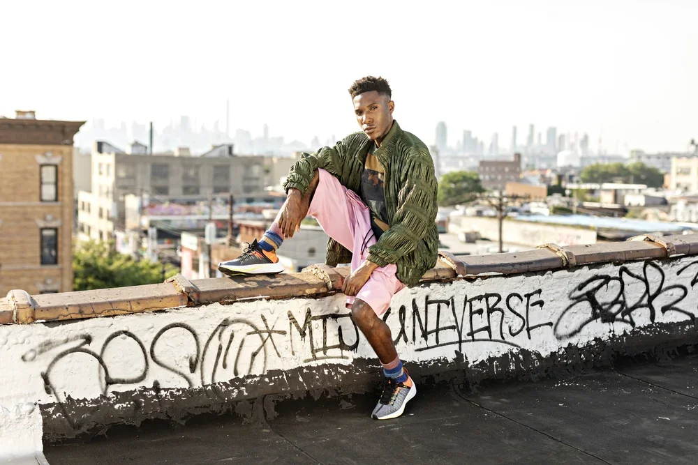 A young man wearing a green jacket, pink shorts, and sneakers, sitting on a graffiti-covered rooftop ledge with an urban cityscape in the background.