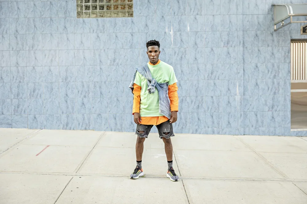 A young man standing alone on a sidewalk in front of a blue tiled wall with a small window and an open gate. He is wearing a layered outfit with bright green, orange, and grey colors, shorts, sneakers, and socks, with a grey jacket draped over his sh
