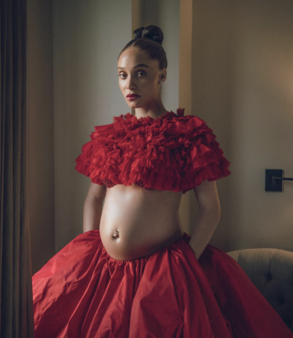 A woman wearing a dramatic red ruffled top and a matching voluminous skirt, standing in a room with neutral-colored walls and furniture.