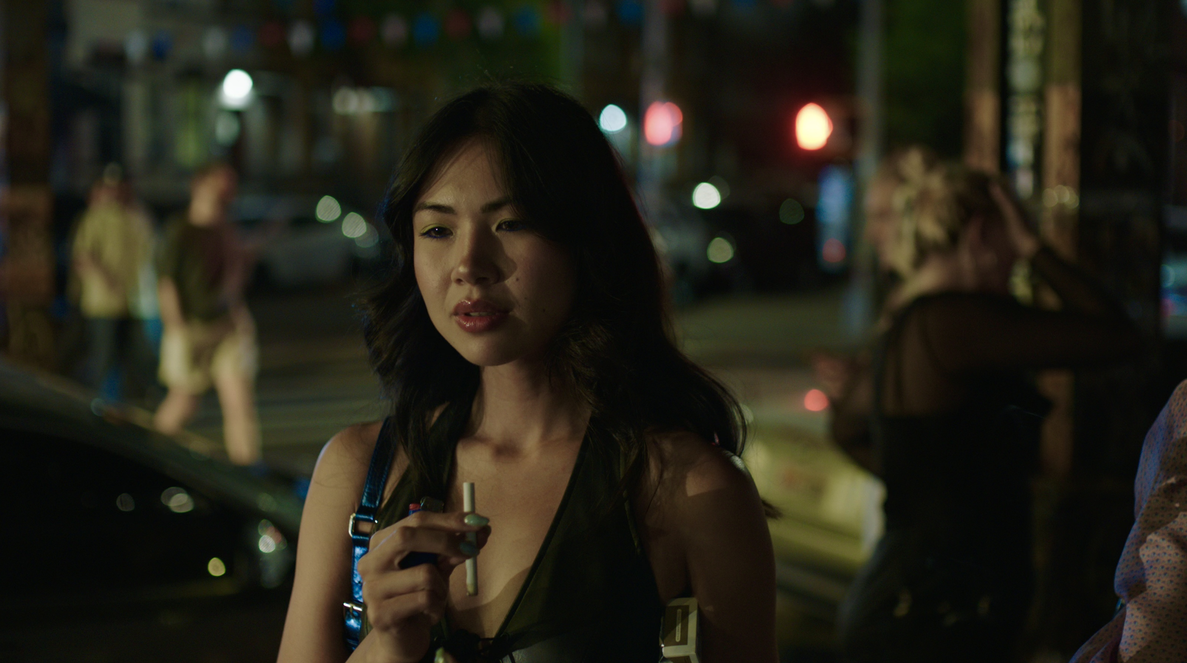 A woman with dark hair looking slightly to the side at night, holding a vape, with blurred colorful city lights in the background.