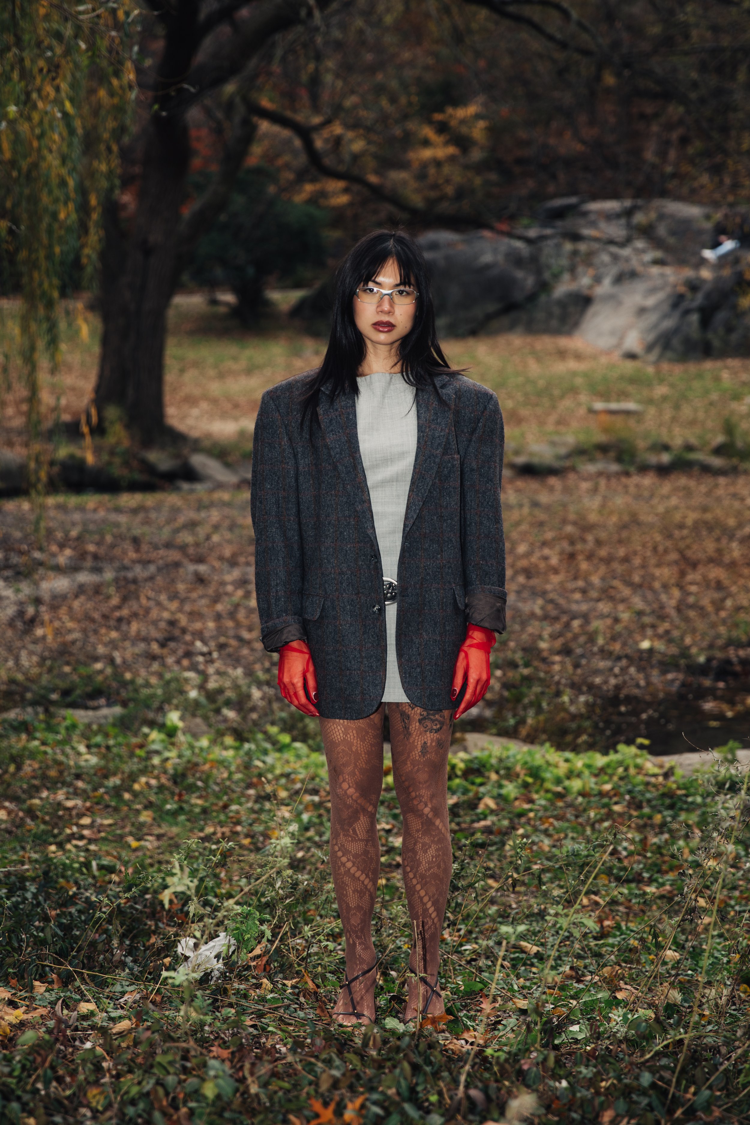 A woman standing outdoors in a park with fall foliage, wearing a plaid blazer, lace tights, red gloves, glasses, and dark lipstick.