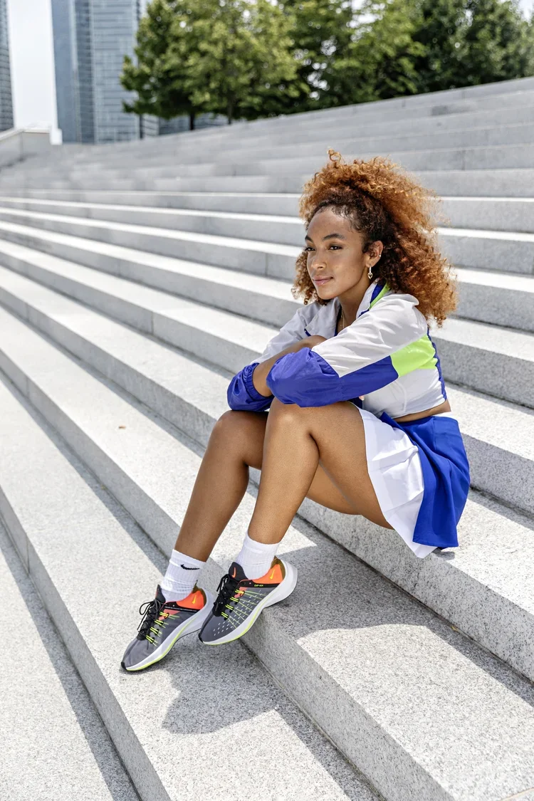 A young woman with curly reddish-brown hair sitting on outdoor stairs, wearing athletic clothes including a white and blue windbreaker, white shorts, and running shoes, with a cityscape and trees in the background.