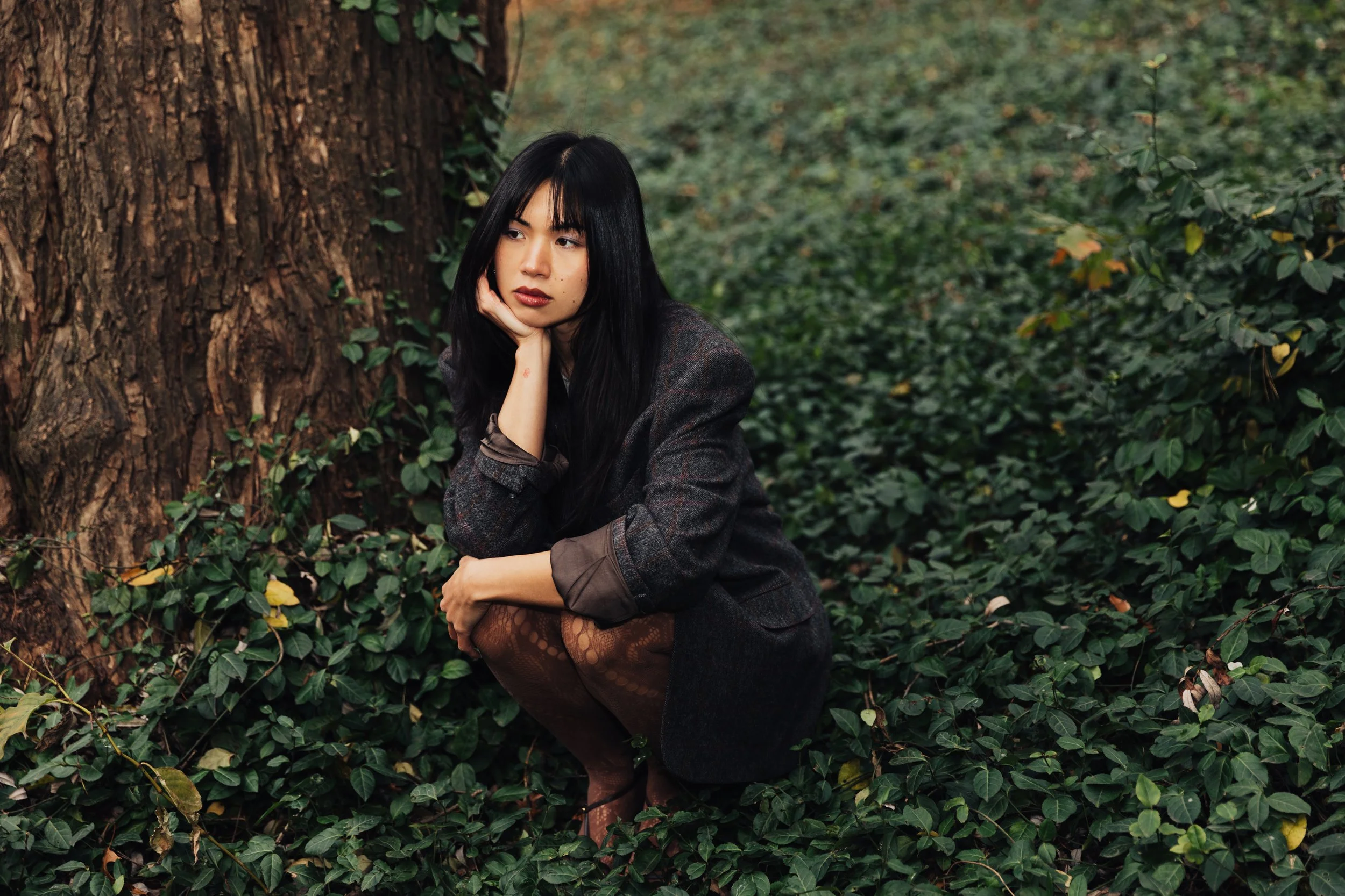 A young woman with long dark hair in a plaid blazer, sitting outdoors beside a large tree with lush green foliage in the background, resting her chin on her hand and looking thoughtfully to the side.