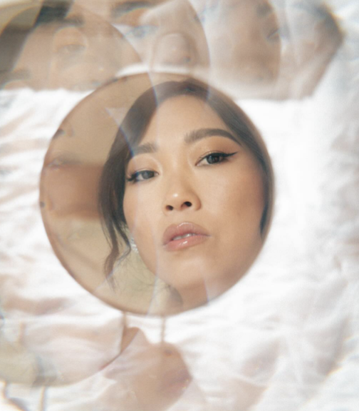 A woman looking into a handheld mirror, with a distorted, layered reflection of her face above her.