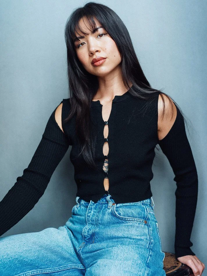 A young woman with long dark hair and bangs, wearing a black cold-shoulder top with button details and light blue jeans, sitting against a plain gray wall.
