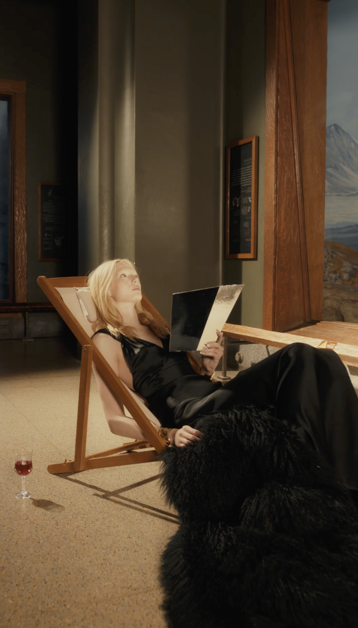 A woman in a black dress relaxing on a lounge chair in a museum, reading a book, with a glass of red wine on the floor nearby.