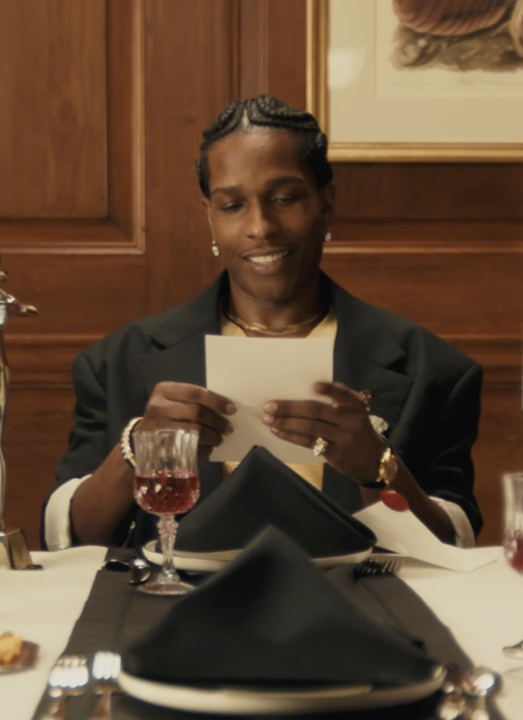 A woman with braided hair wearing earrings, a necklace, and a black blazer sitting at a dinner table, holding and reading a card with a smile.