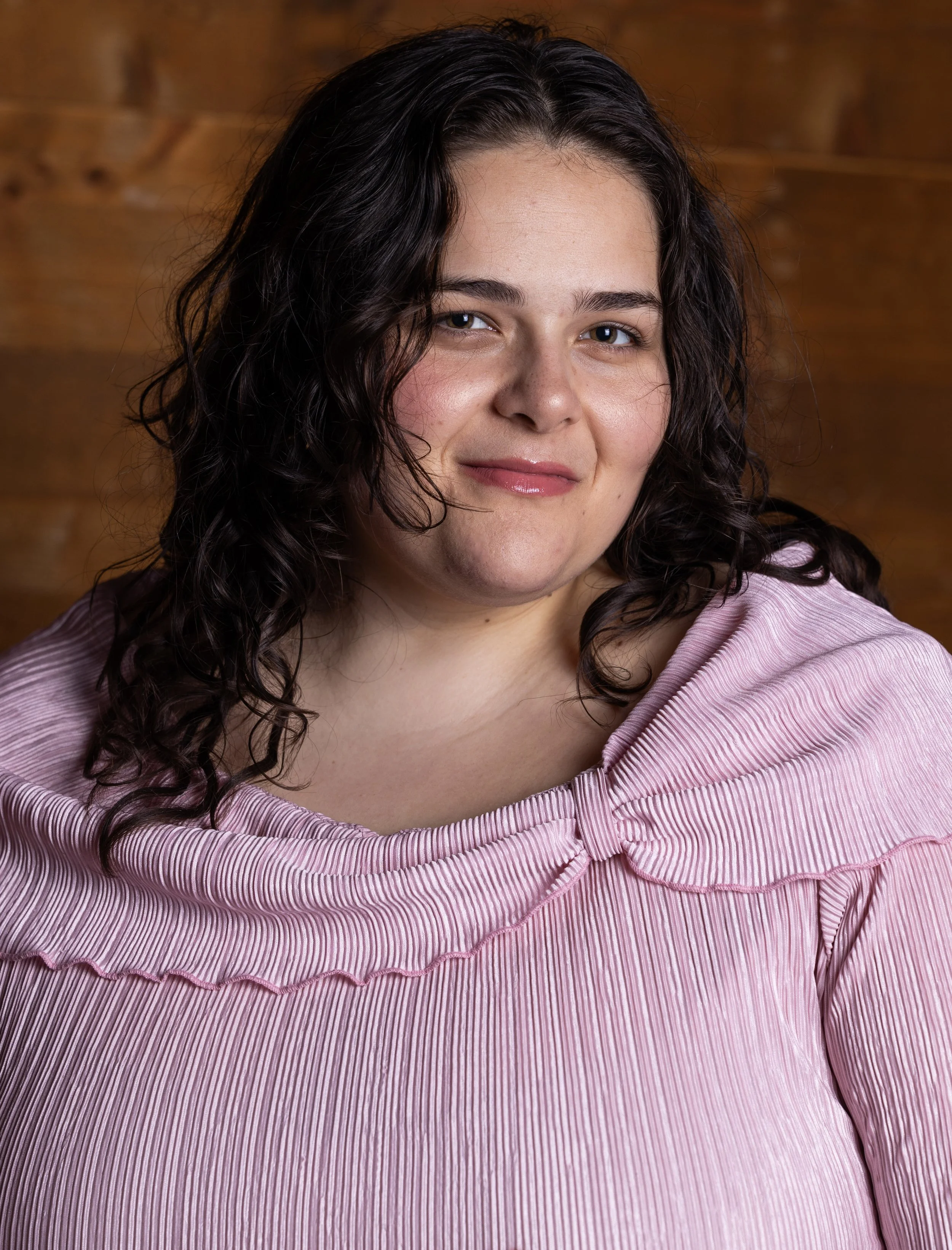 A woman with dark, curly hair and fair skin wearing a pink, pleated, ruffled top.