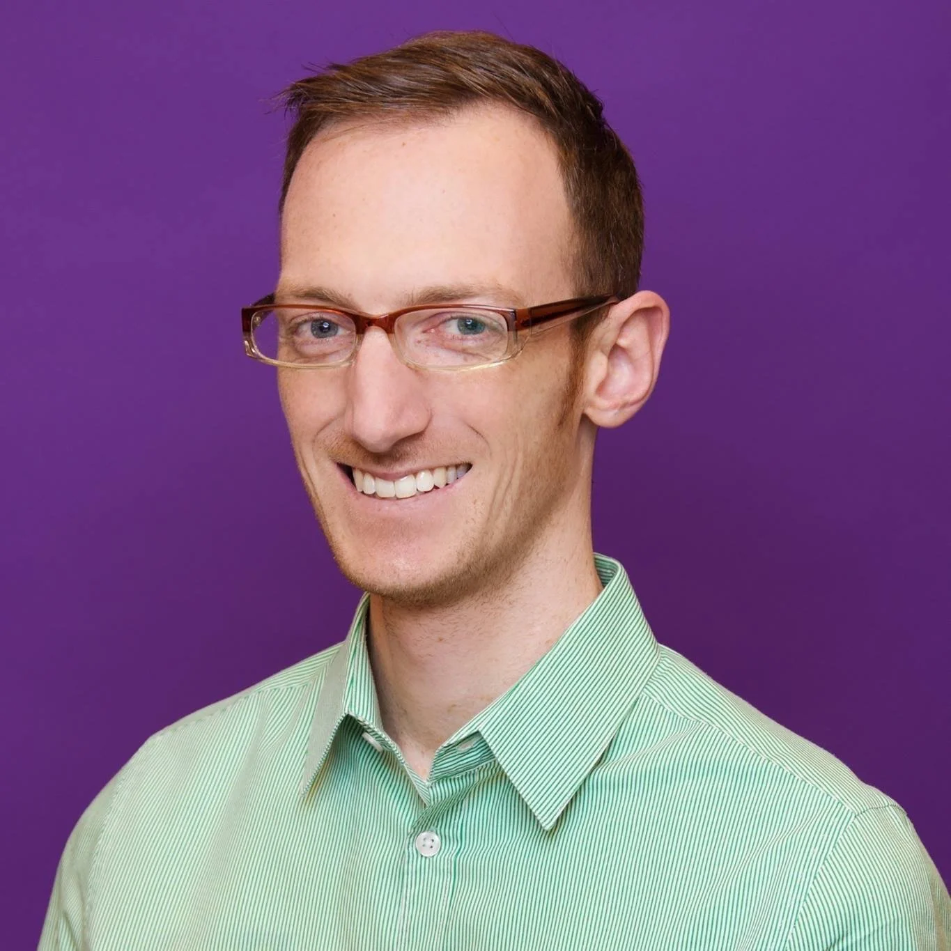 A man with short brown hair, glasses, and a big smile, wearing a light green striped shirt, standing in front of a purple background.