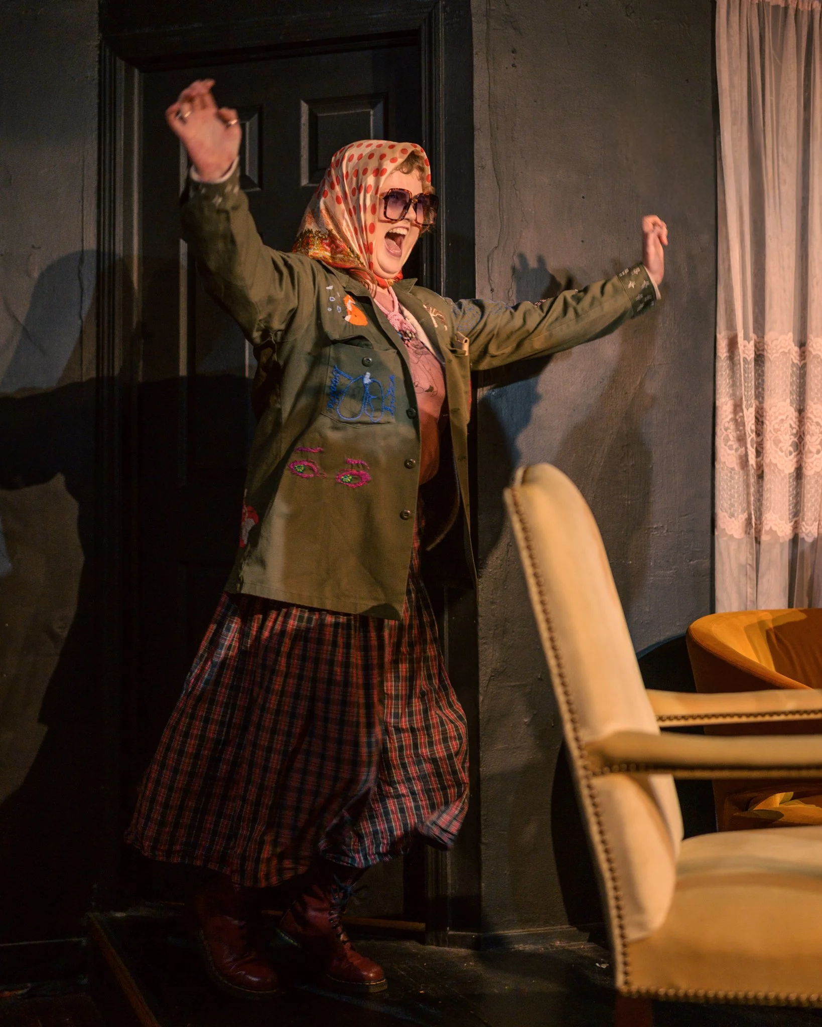 Woman with curly hair wearing large sunglasses, a polka dot headscarf, and a green jacket with colorful patches, standing with arms outstretched and shouting in front of a dark wall with a door, next to a beige chair with a pom-pom trim in a room with dark decor.