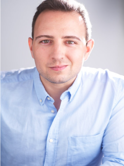 Headshot of a young man with short dark hair, wearing a light blue button-up shirt, smiling softly against a plain background.