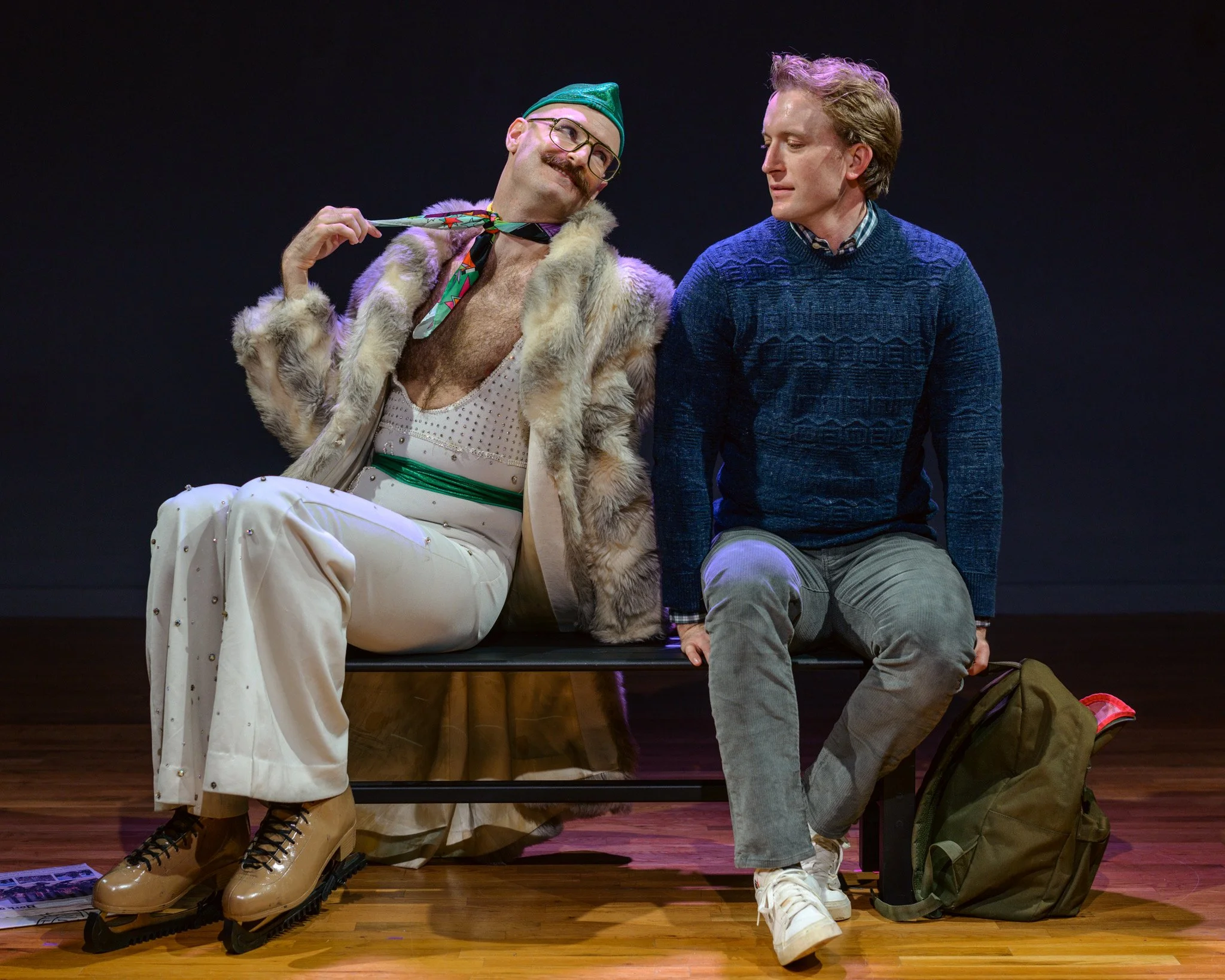 Two men sitting on a stage, one dressed as a clown or performer with a furry coat, glasses, a green hat, and beige boots, and the other in casual clothing with a blue sweater and gray pants, engaged in conversation or performance.