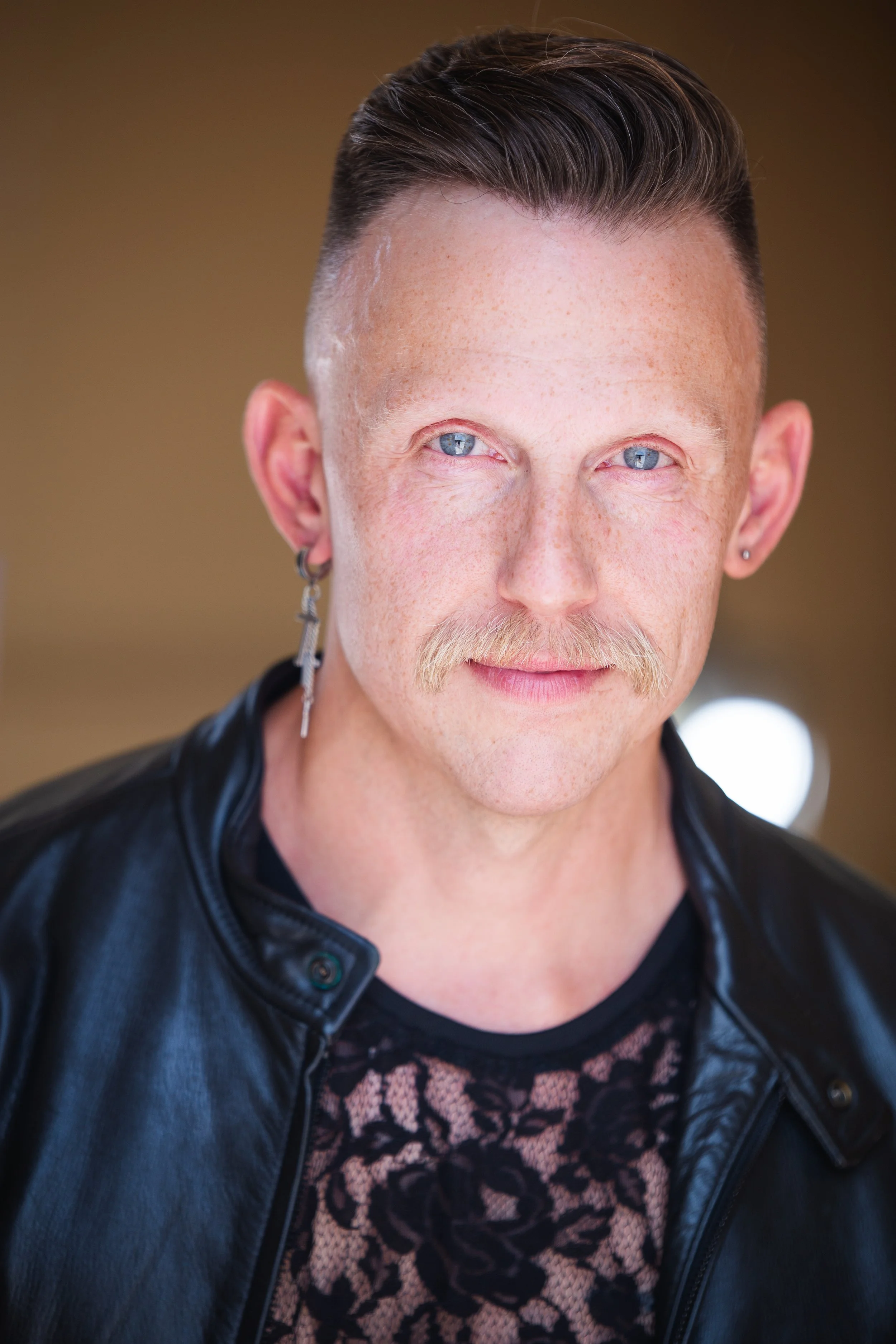 A close-up portrait of a man with dyed blue eyes, light skin, a mustache, and multiple earrings, wearing a black leather jacket and a leopard print shirt, against a blurred background.