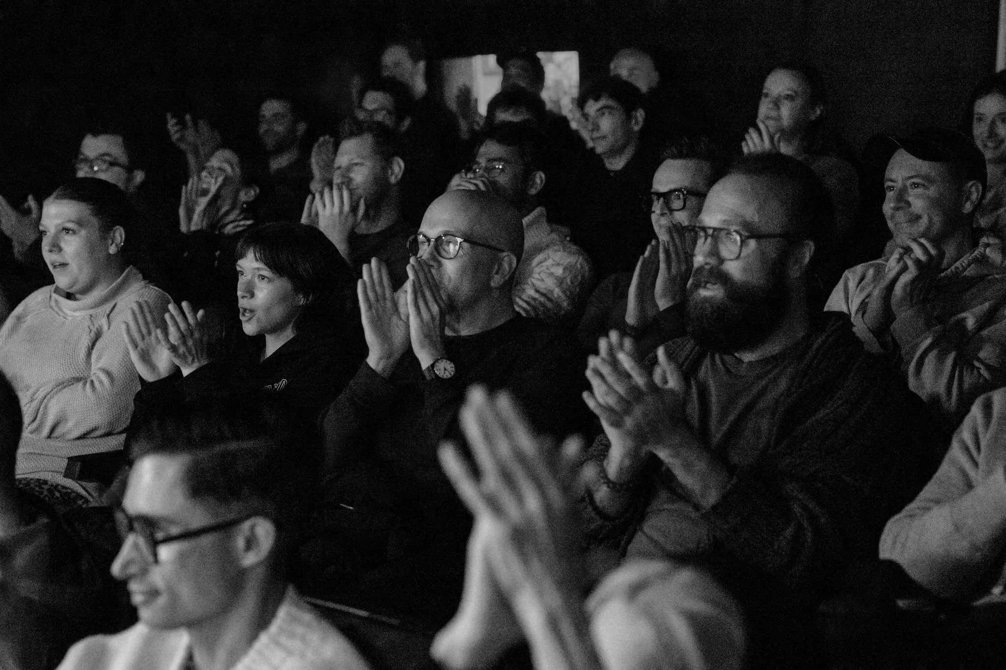 People seated in an audience watching an event, some are laughing and smiling, in black and white.