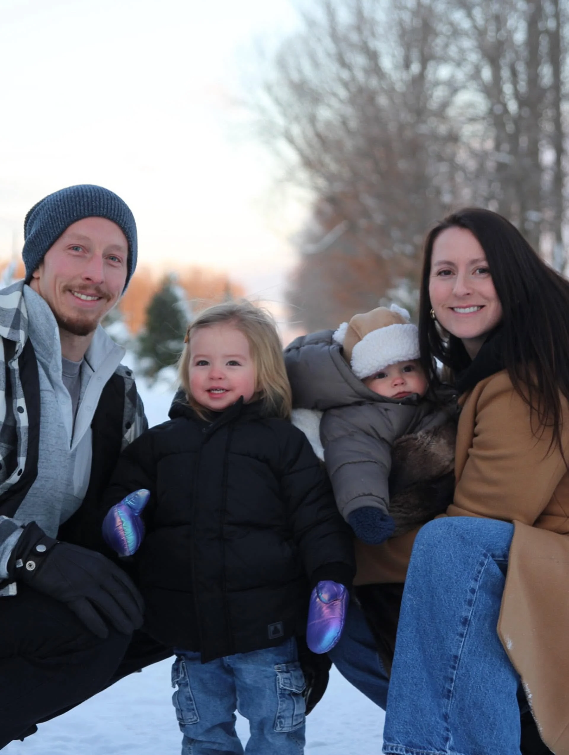 A family of five, including two adults and three children, smiling outdoors in winter, wearing warm clothing, with snow and leafless trees in the background.