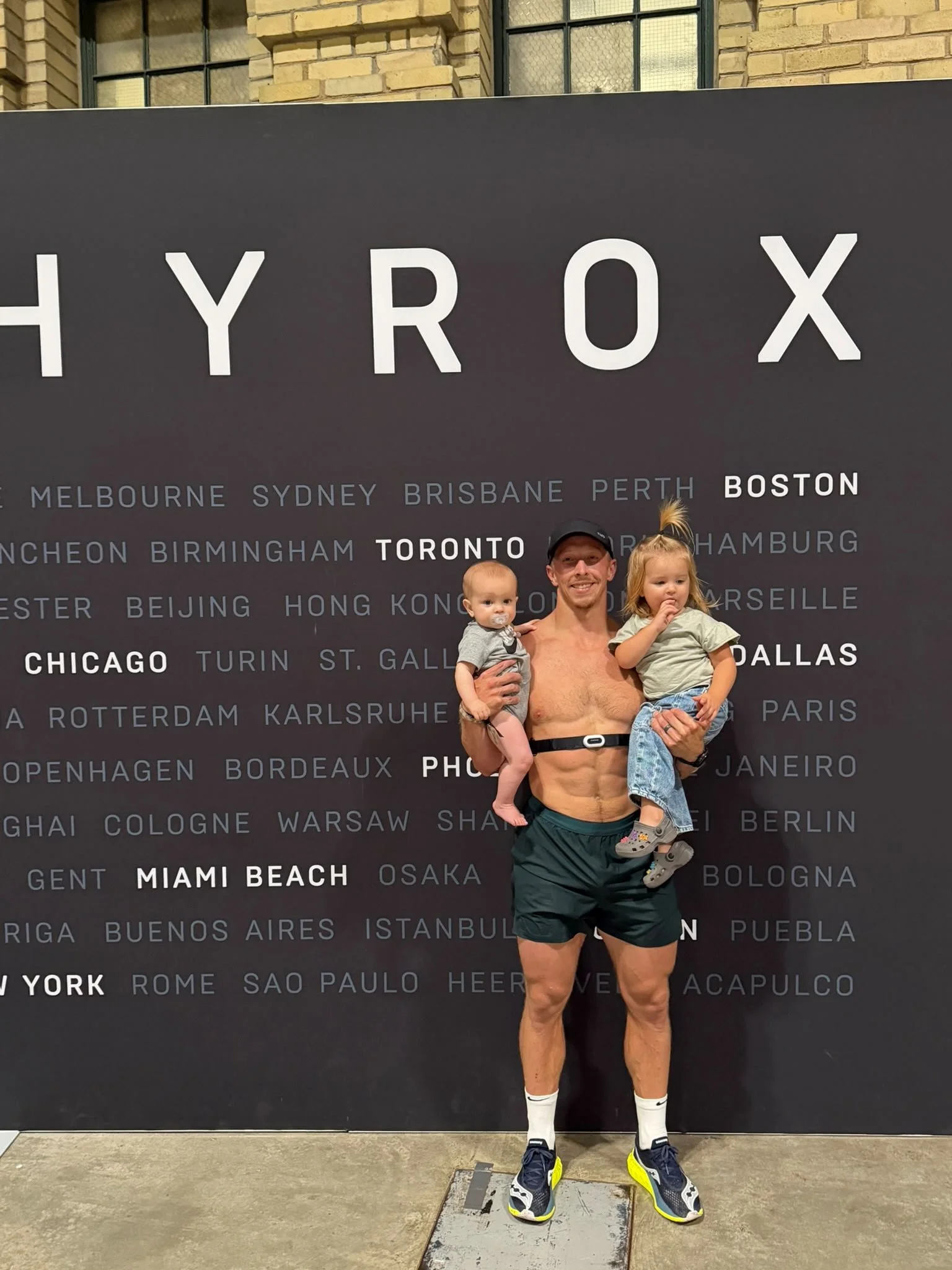 A man holding two children in front of a black wall with city names. The man is shirtless, wearing shorts, sneakers, and socks. The children are wearing casual clothing. The background features a brick building and a sign listing various city names.