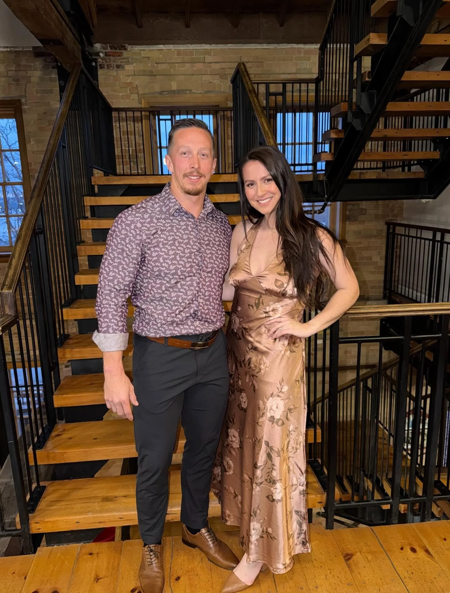 A man and woman standing on a wooden staircase inside a brick building. The man has short slicked-back hair, a beard, and is wearing a patterned shirt with rolled-up sleeves, dark pants, and brown shoes. The woman has long dark hair, and is wearing a